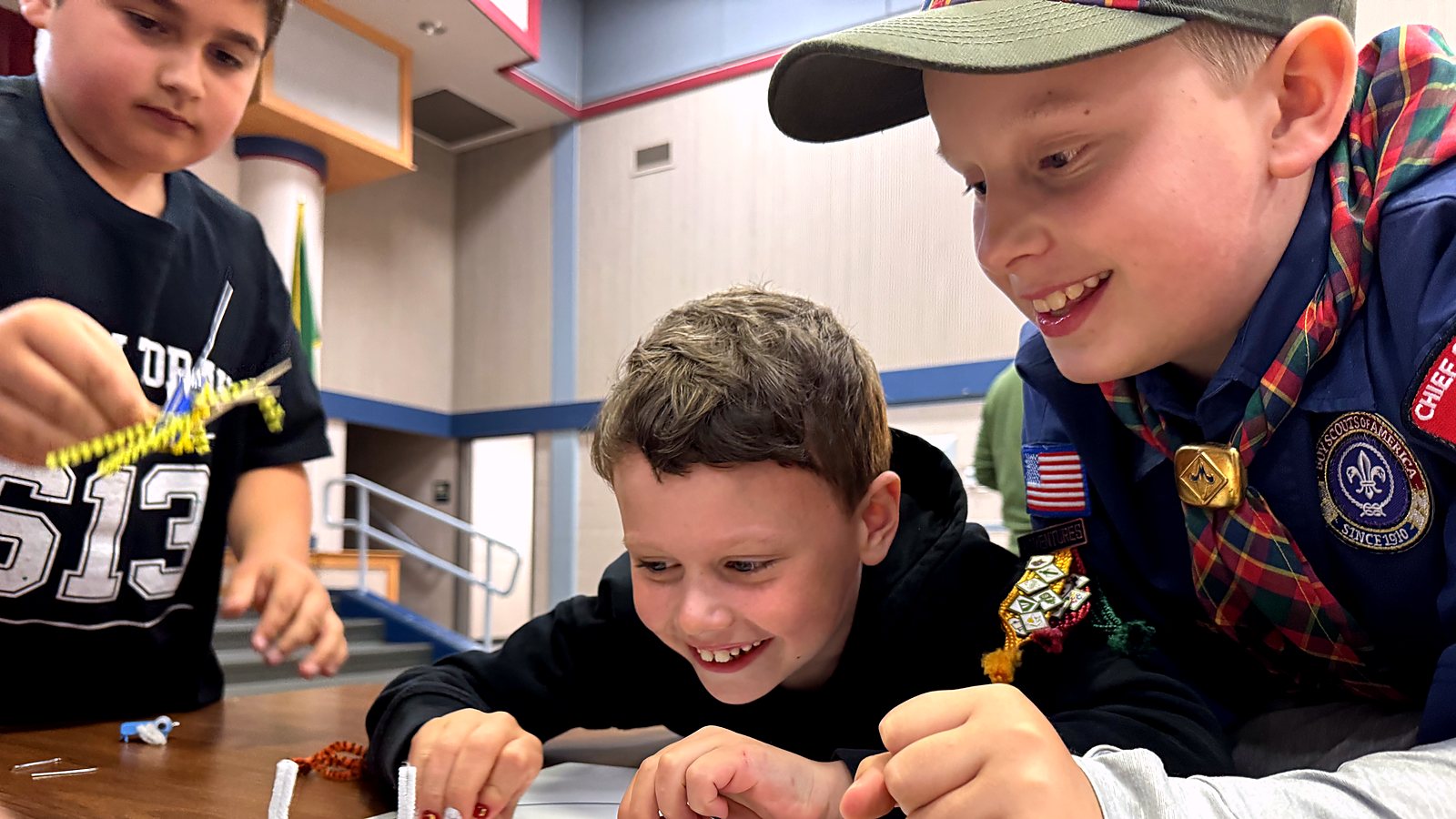 Three Cub Scouts leaning over a table, grinning, building something out of pipe cleaners at a pack meeting.